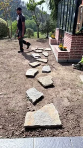 Operario colocando sendero de losas de piedra natural irregular sobre suelo sin césped junto al invernadero victoriano de aluminio negro en Errigoiti (Bizkaia)