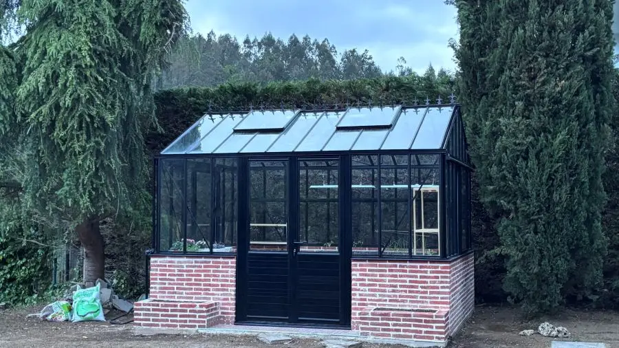 Vista frontal de invernadero victoriano de aluminio negro con cubierta de vidrio a dos aguas y ventanas abatibles.