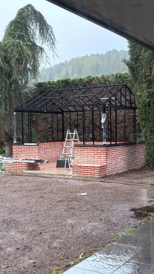 Vista frontal de la estructura de aluminio negro de un invernadero victoriano sin acristalar sobre zócalo de ladrillo visto rojo, fotografiada bajo la lluvia desde el interior de una terraza cubierta.